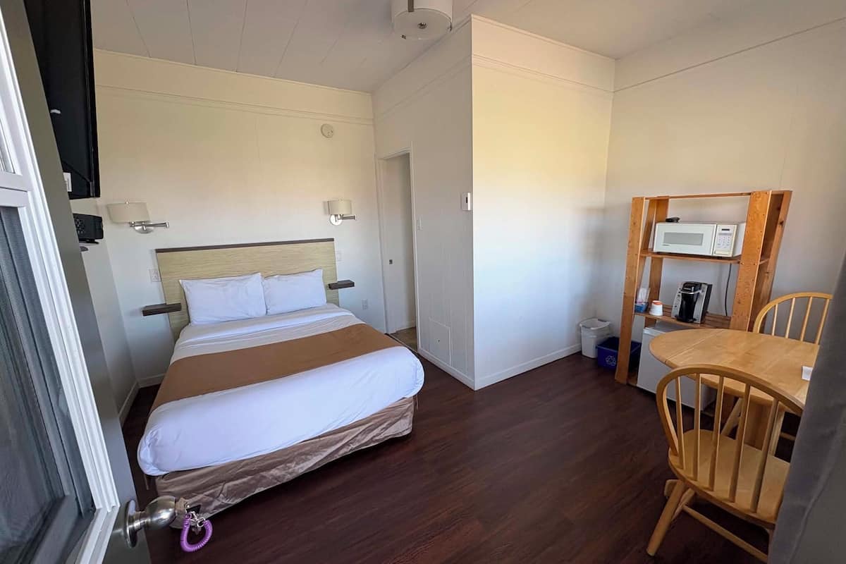 Habitación de hotel con cama, una silla de madera, una mesa con microondas y un cubo de basura pequeño.