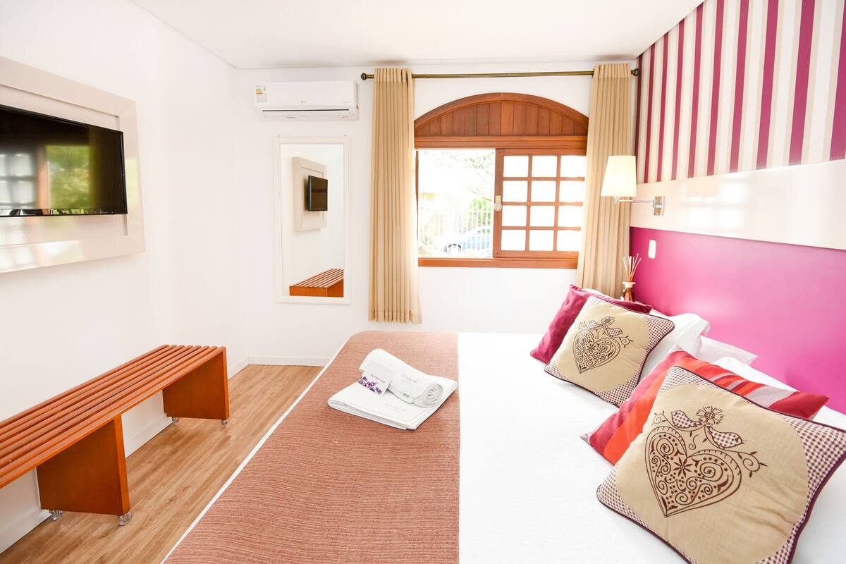 A hotel room with a bed, a bench, a TV, and a window with curtains.