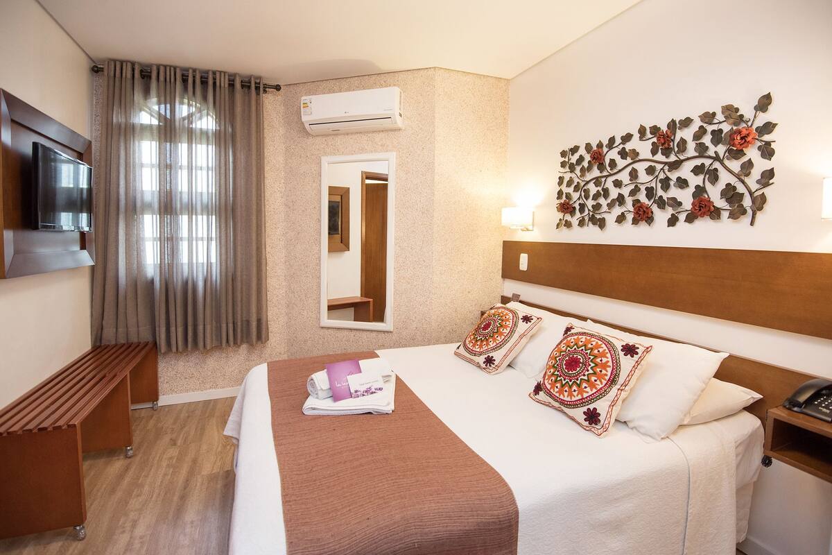 A hotel room with a bed, a TV, an air conditioner, a mirror, a bedside table, and a decorative wall piece.