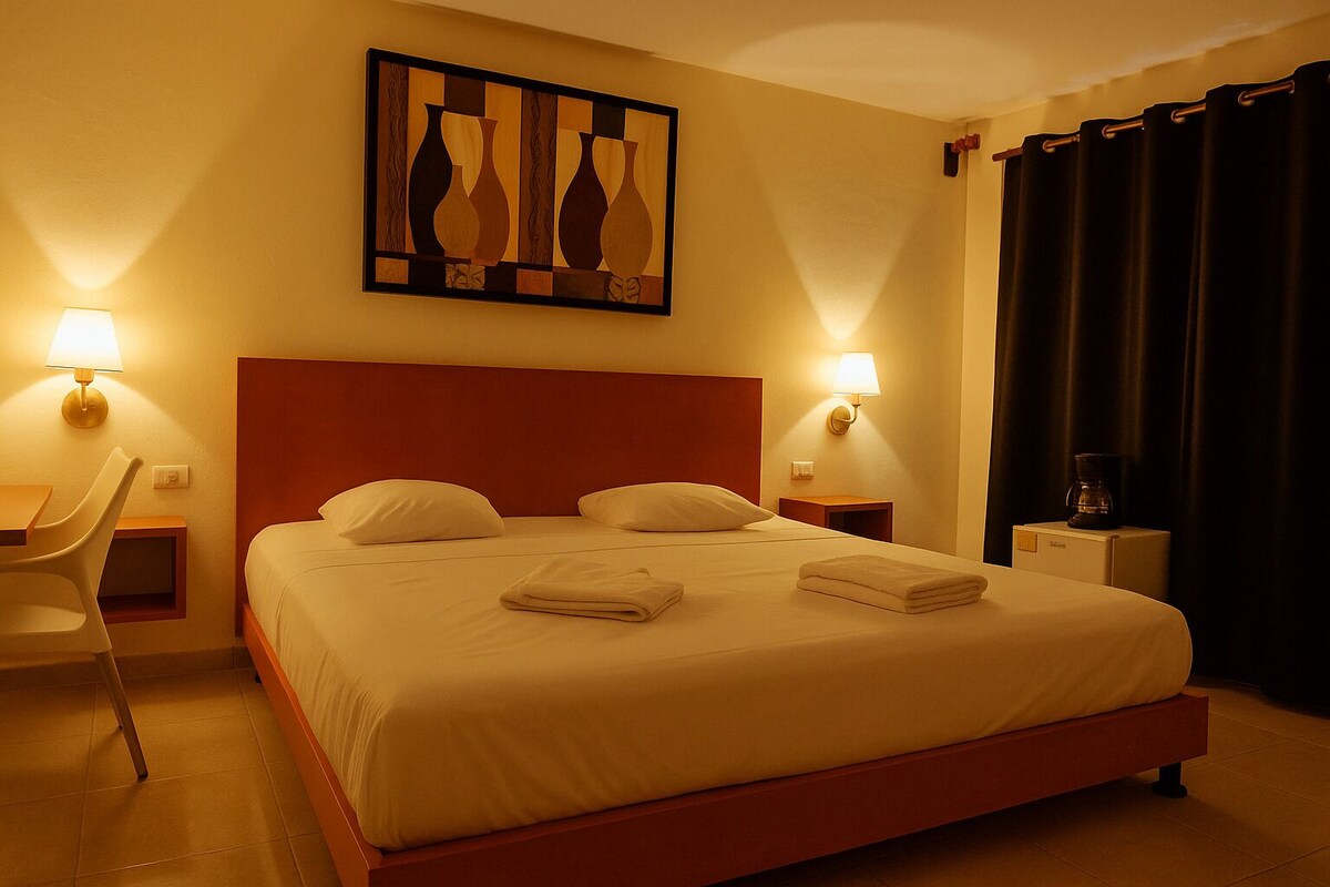 A hotel room with a bed, two bedside lamps, a desk, a chair, and a painting on the wall.