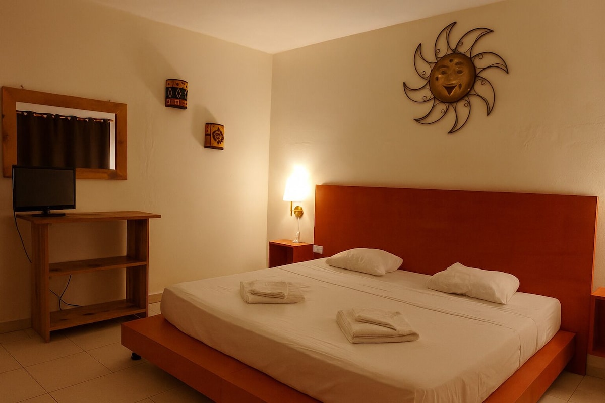 A hotel room with a bed, a TV on a wooden stand, a wall-mounted sun decoration, and a nightstand with a lamp.