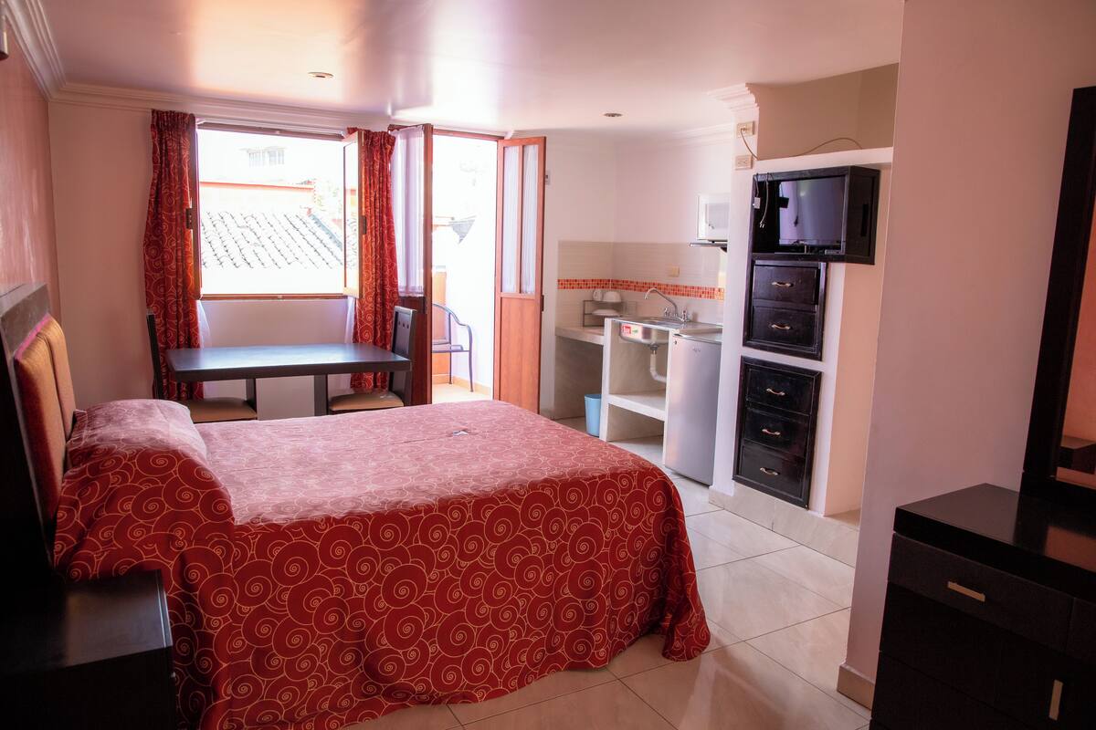 A hotel room with a bed, a desk, a chair, a small kitchenette, and a TV.