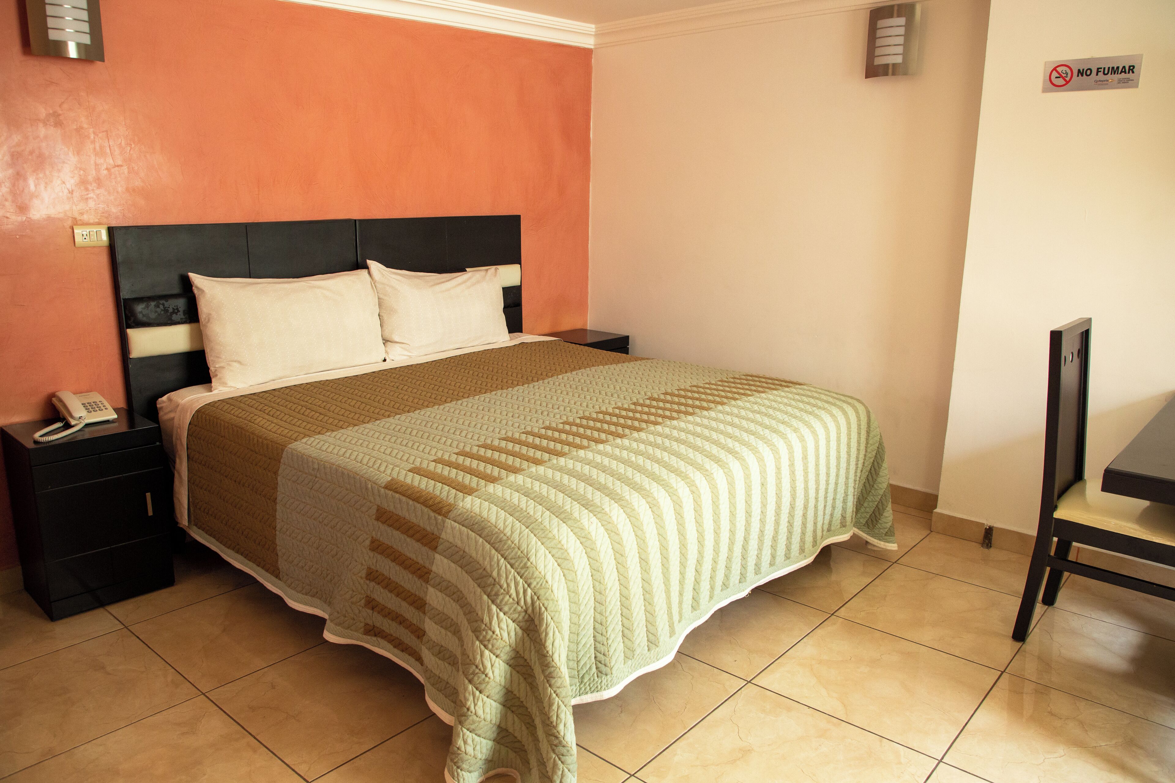 A neatly made bed with a striped blanket, a bedside table with a phone, and a chair in a room with orange walls and a tiled floor.