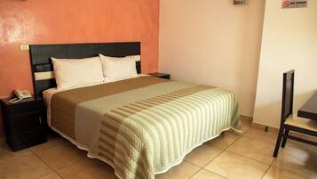 A neatly made bed with a striped blanket, a bedside table with a phone, and a chair in a room with orange walls and a tiled floor.
