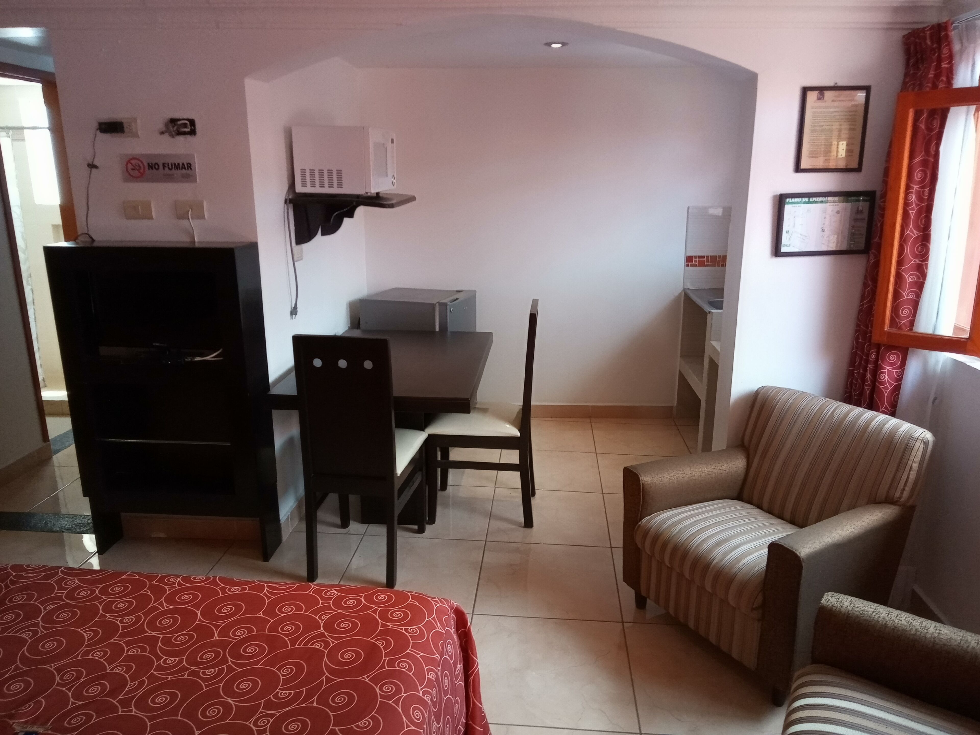 A hotel room with a bed, a desk with a chair, a small kitchenette, and a TV.