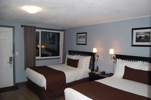 A hotel room with two beds, a window with curtains, a framed picture on the wall, and a nightstand with a lamp.