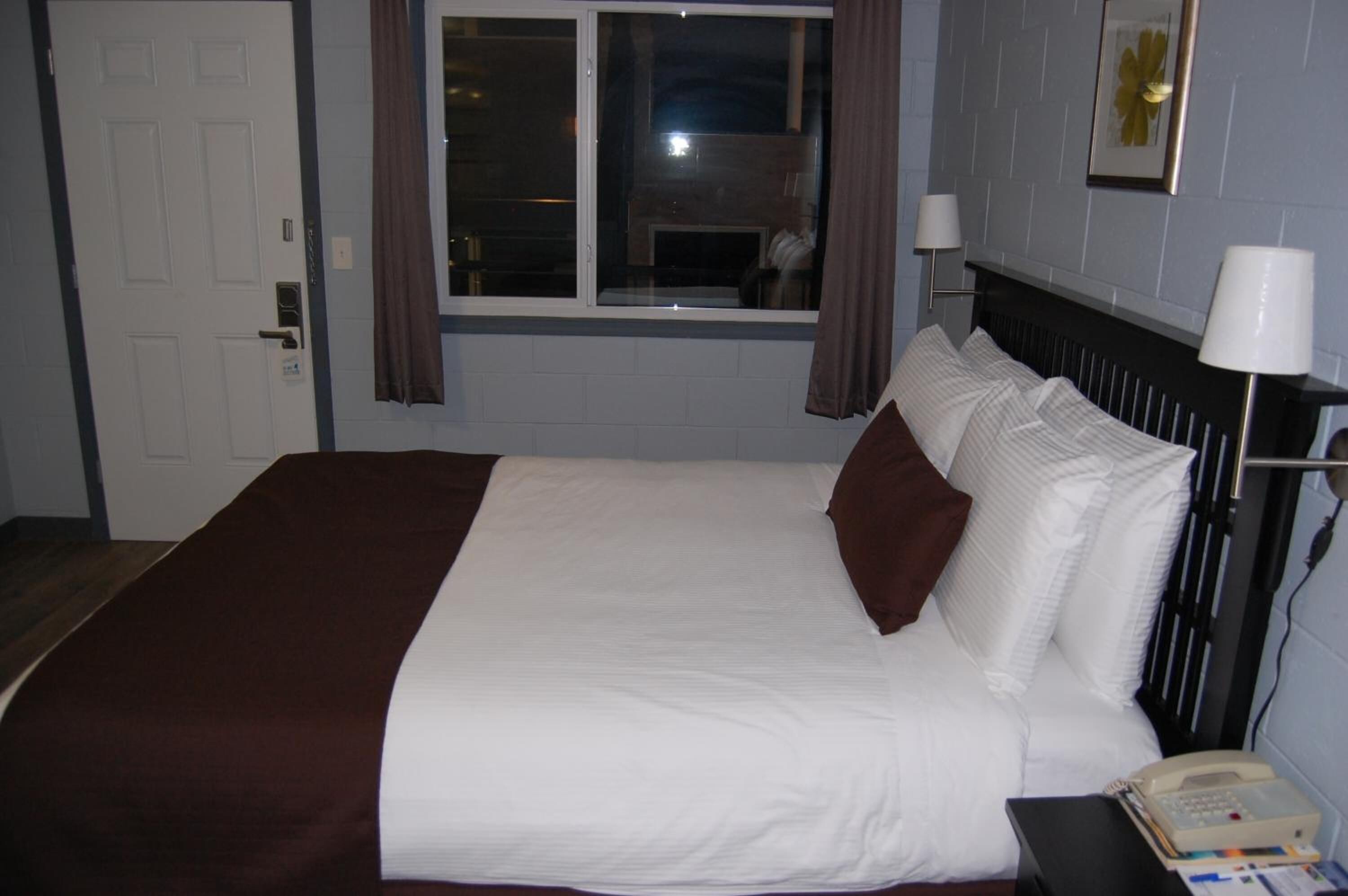 A hotel room with a bed, a window, a door, and a bedside table with a lamp.