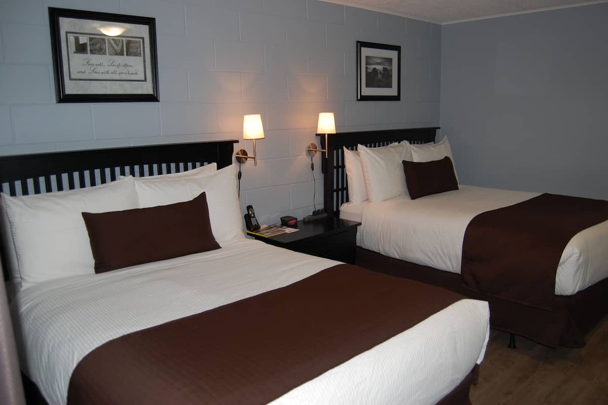 Ein Hotelzimmer mit zwei Betten, jeweils mit weißen Laken und braunen Zierkissen. An jeder Seite der Betten befinden sich Wandlampen und über dem linken Bett ein gerahmtes Bild.