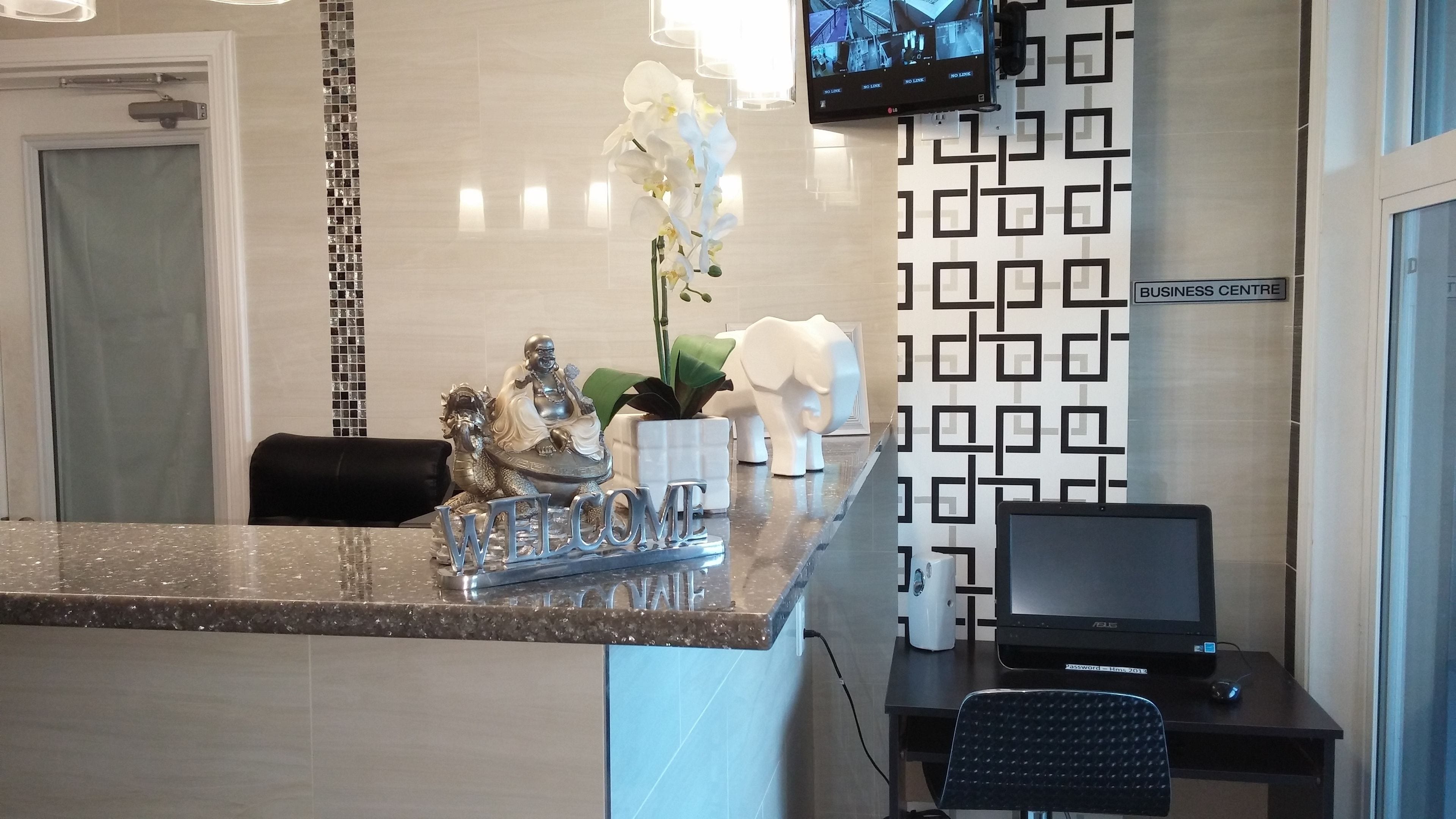 A reception area with a marble counter, a computer, a monitor, a black chair, a TV mounted on the wall, and a sign indicating the business center.