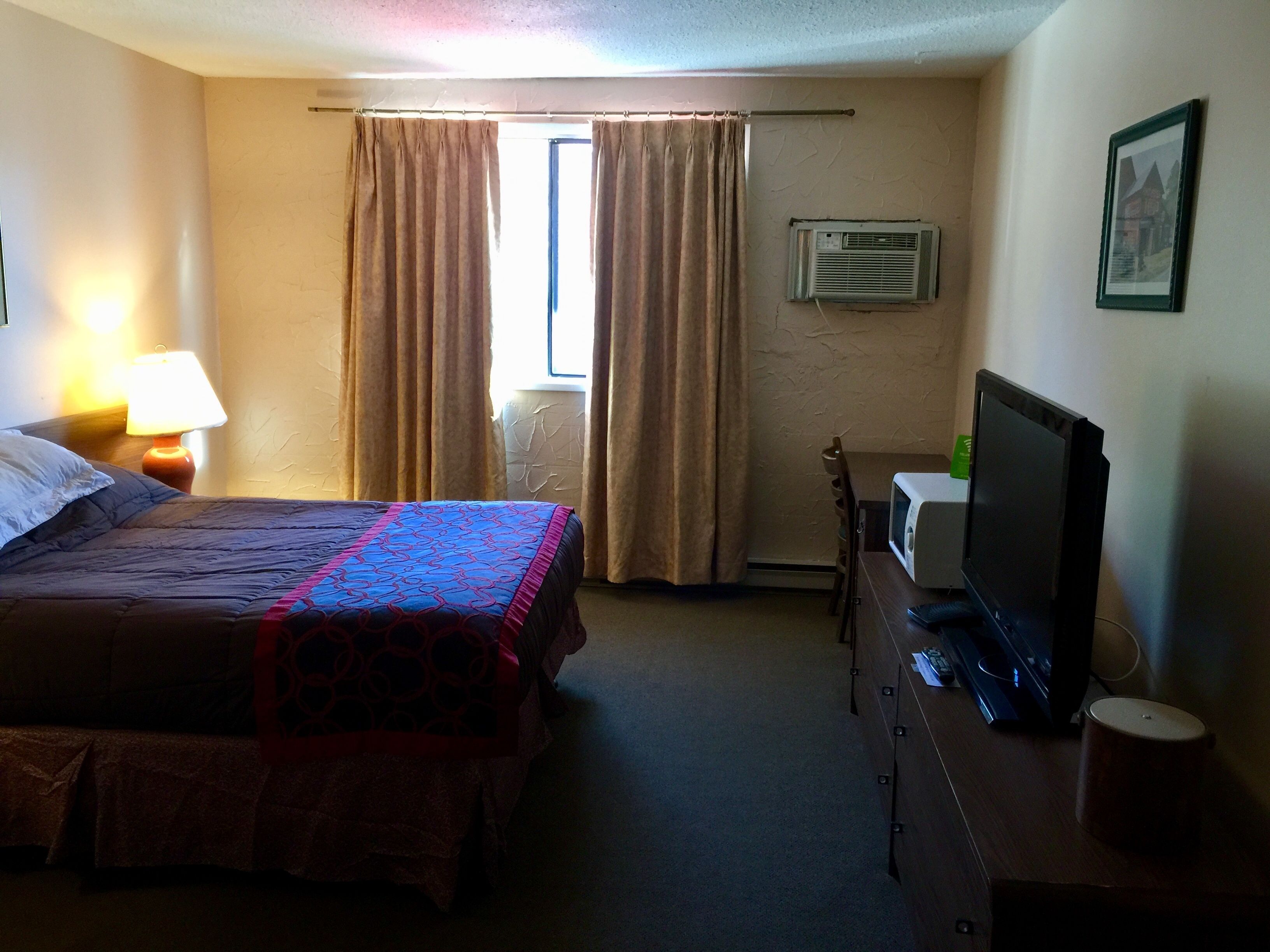 A hotel room with a bed, a desk with a television, a lamp, and a window with curtains.