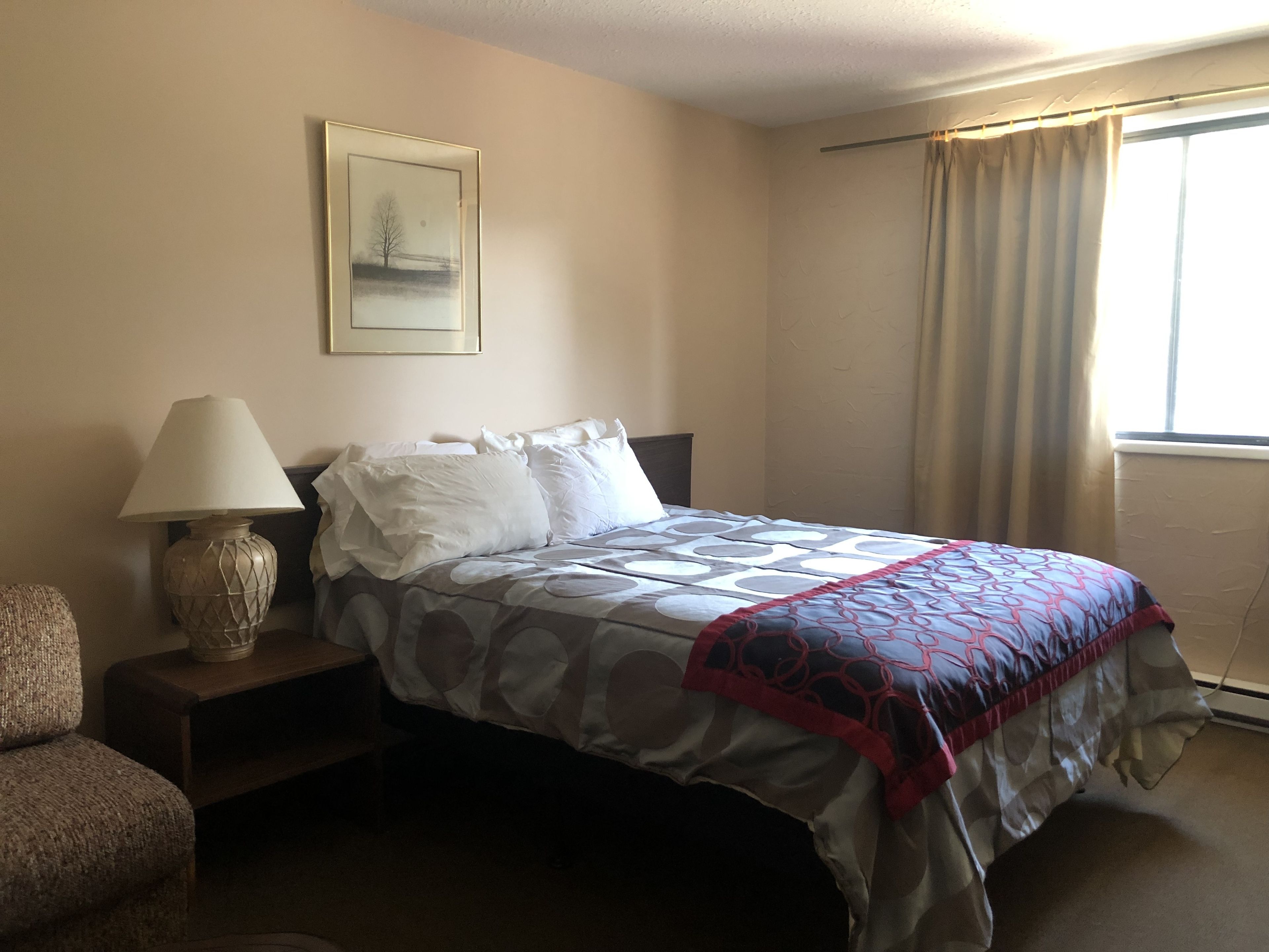 Une chambre d’hôtel bien rangée, avec un lit, une lampe de chevet, une chaise et une fenêtre avec des rideaux.