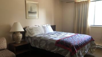 Une chambre d’hôtel bien rangée, avec un lit, une lampe de chevet, une chaise et une fenêtre avec des rideaux.