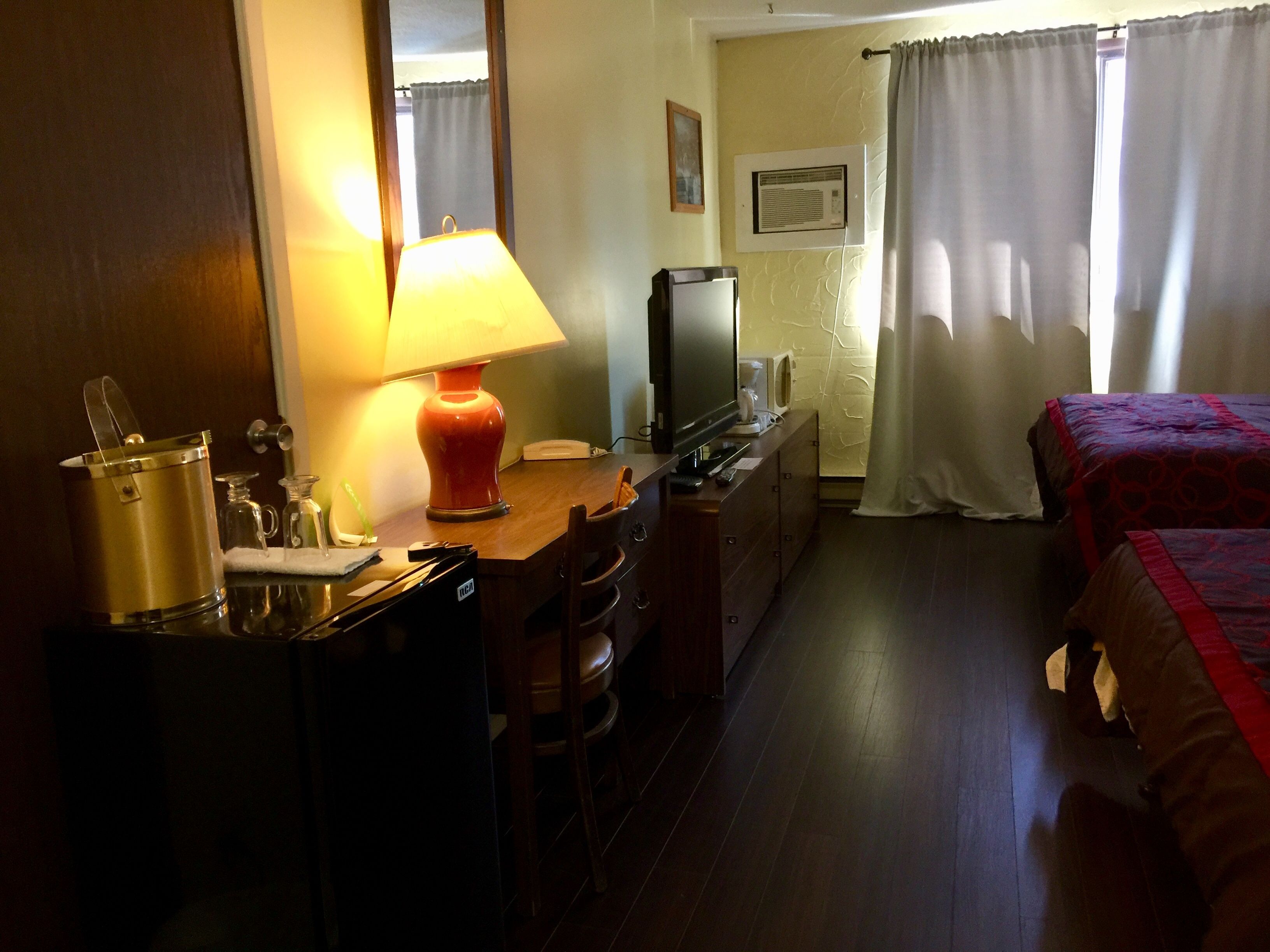 Habitación de hotel con un escritorio de madera, una lámpara, un televisor, una cama con manta roja y una ventana con cortinas.