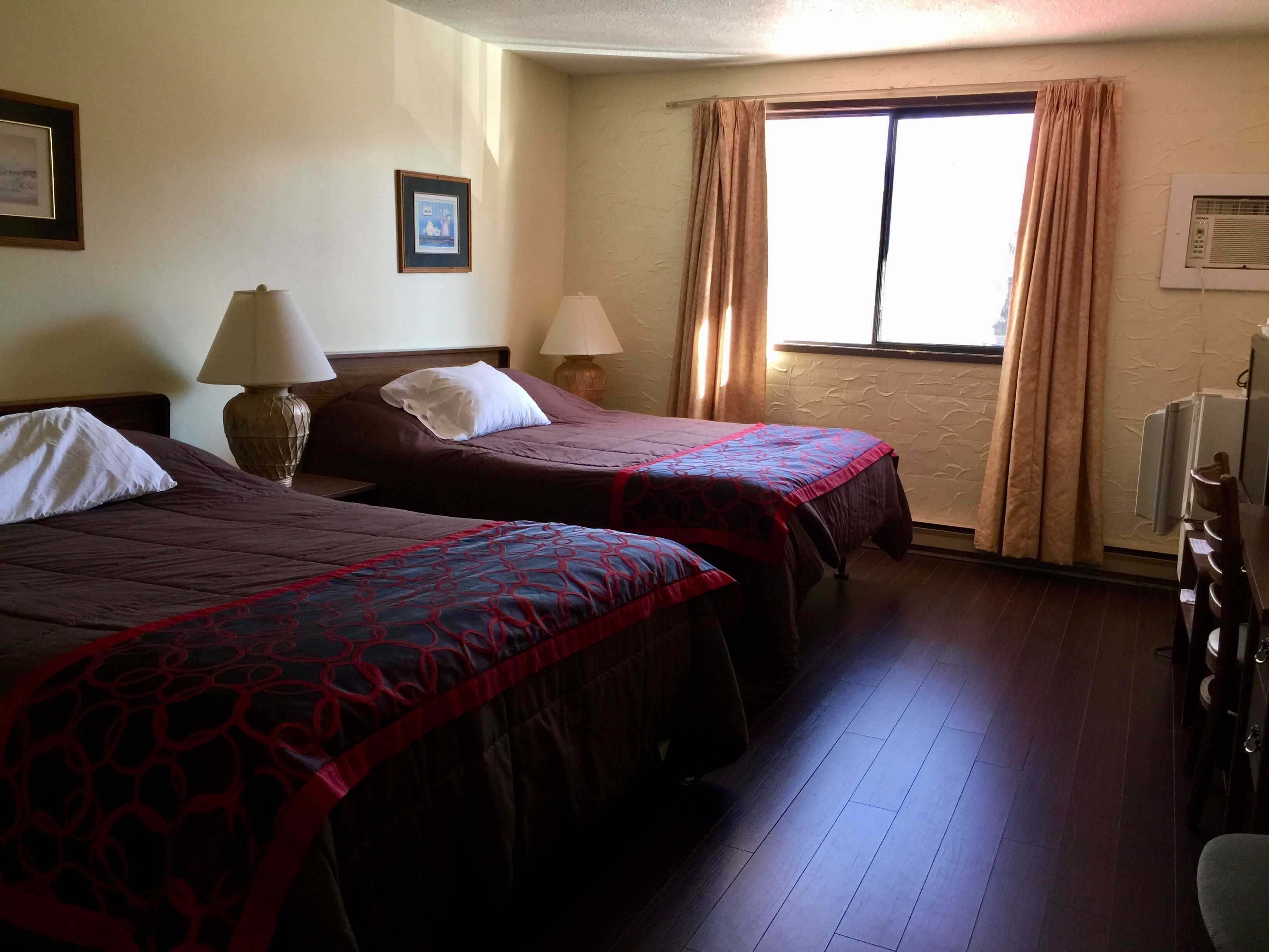 A hotel room with two beds, a window with curtains, a lamp, and a radiator.