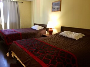 A hotel room with two beds, each with a brown bedspread and red circular patterns. There is a lamp on the nightstand between the beds, and a painting on the wall above the beds.