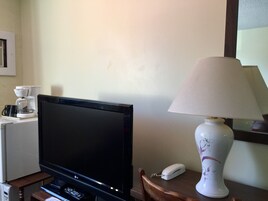 Un televisor sobre un soporte, una cafetera, una lámpara y un teléfono sobre una mesa de madera en una habitación con pared lisa.