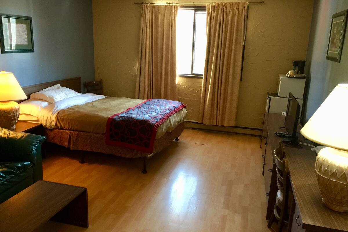 A hotel room with a bed, a desk, a chair, a lamp, and a window with curtains.