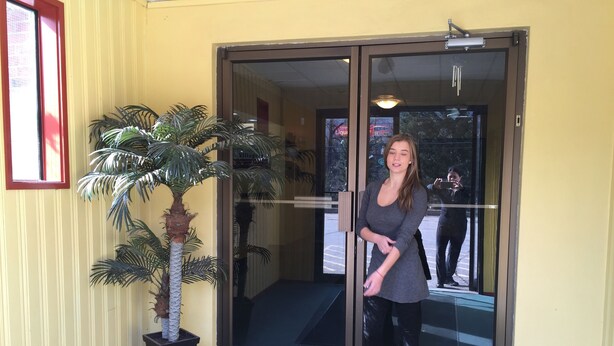 A woman standing at the entrance of a building with a glass door, a potted palm tree, and a red-framed window to the left. - Villager's Inn (Fruitvale)