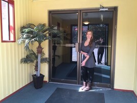 Una mujer de pie frente a la entrada de un edificio con puerta de vidrio, una palmera en maceta y un ventanal de marco rojo a la izquierda.