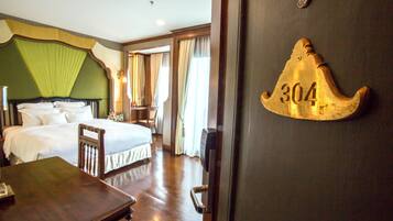 A hotel room with a large bed, a wooden nightstand, a desk, and a door with a number plaque.