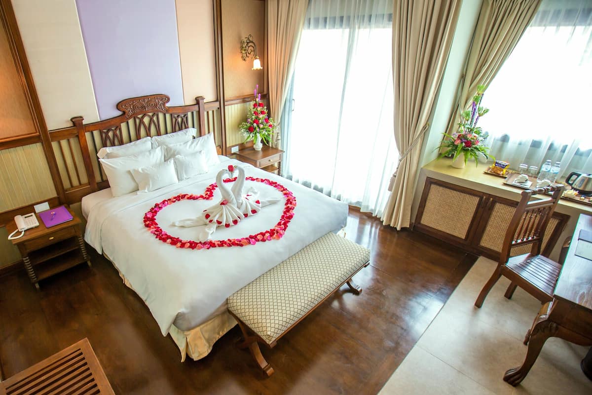 A hotel room with a neatly made bed, a wooden nightstand with a phone and a notepad, a small table with a vase of flowers, and a dining area with a chair and table.