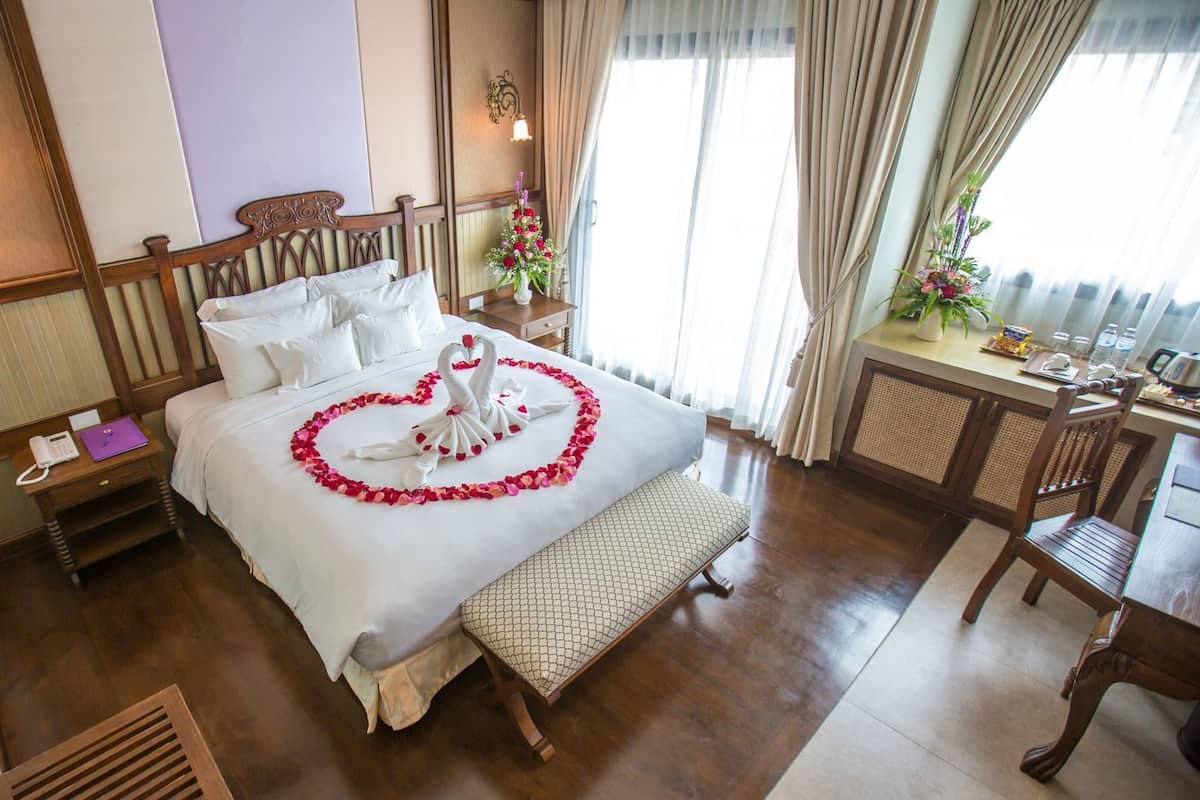 A hotel room with a neatly made bed, a wooden headboard, a bedside table with a phone, a small desk with a chair, and a window with curtains.