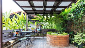 Een terras buiten met een houten terras, metalen stoelen, een ronde bak van baksteen en diverse potplanten.