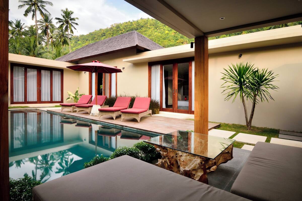 Un área de piscina moderna con sillones de descanso, una mesa de vidrio y vistas a un paisaje tropical.