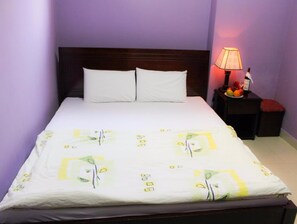 Ein ordentlich bezogenes Bett mit weißer Bettdecke mit Blättermuster, ein hölzerner Kopfteil und ein Nachttisch mit Lampe und Obst.