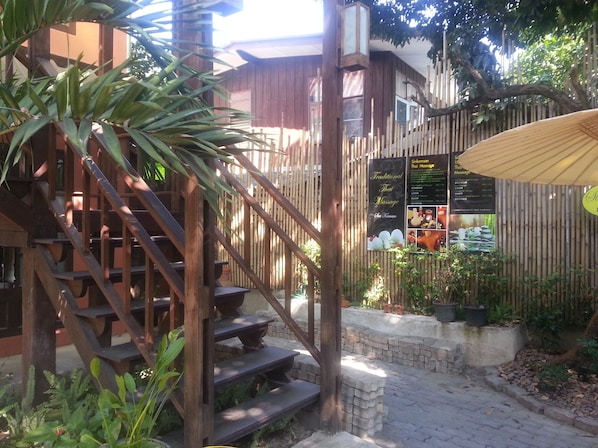 Una escalera de madera conduce a un edificio con cerca de bambú y un letrero que anuncia servicios de masaje tradicional tailandés.