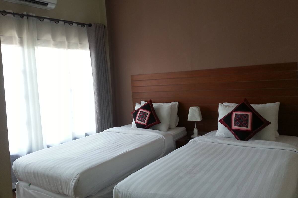 A hotel room with two beds, a wooden headboard, a window with curtains, and an air conditioning unit.