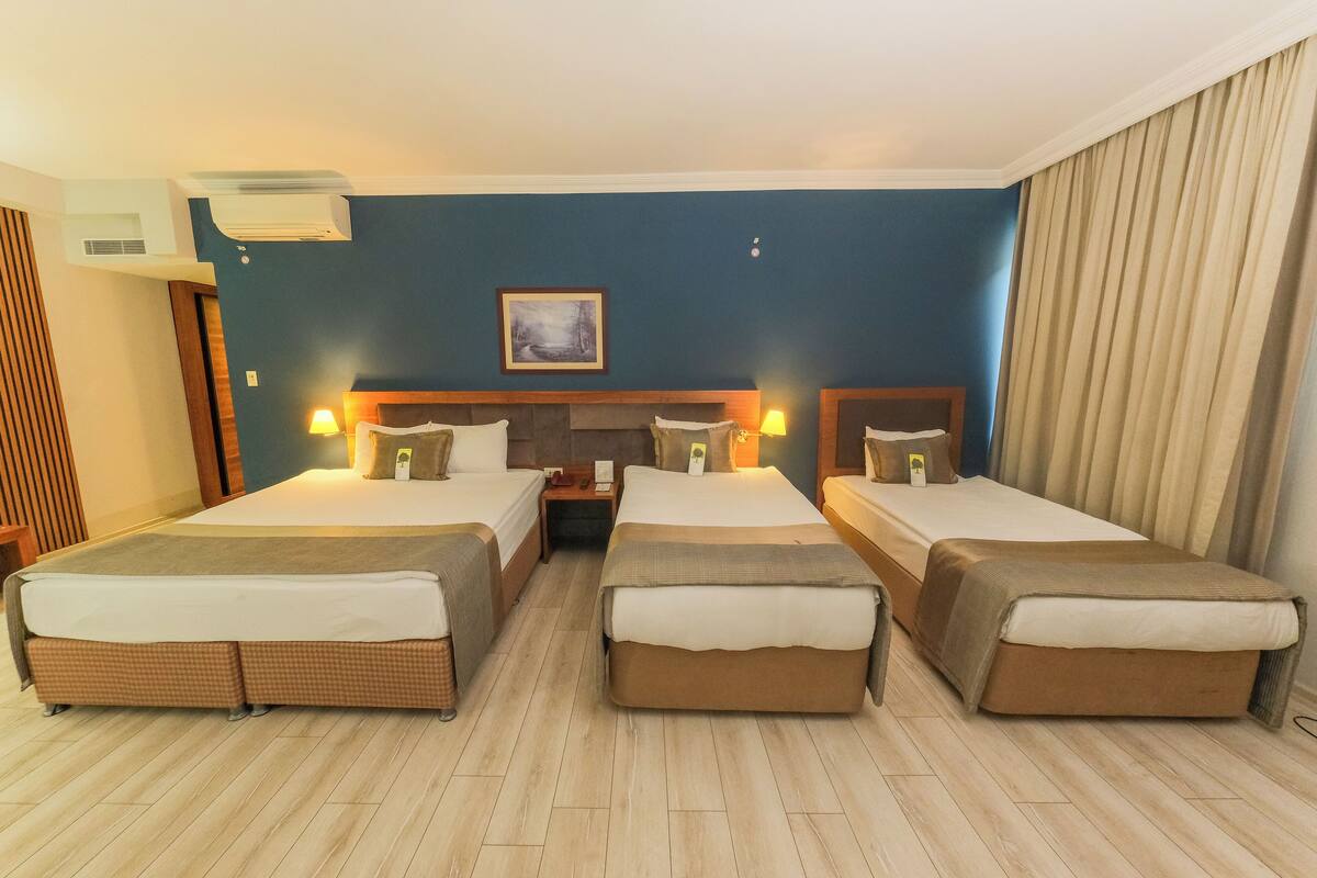 Habitación de hotel con tres camas, cabecera, lámparas de noche, unidad de aire acondicionado y un cuadro en la pared.