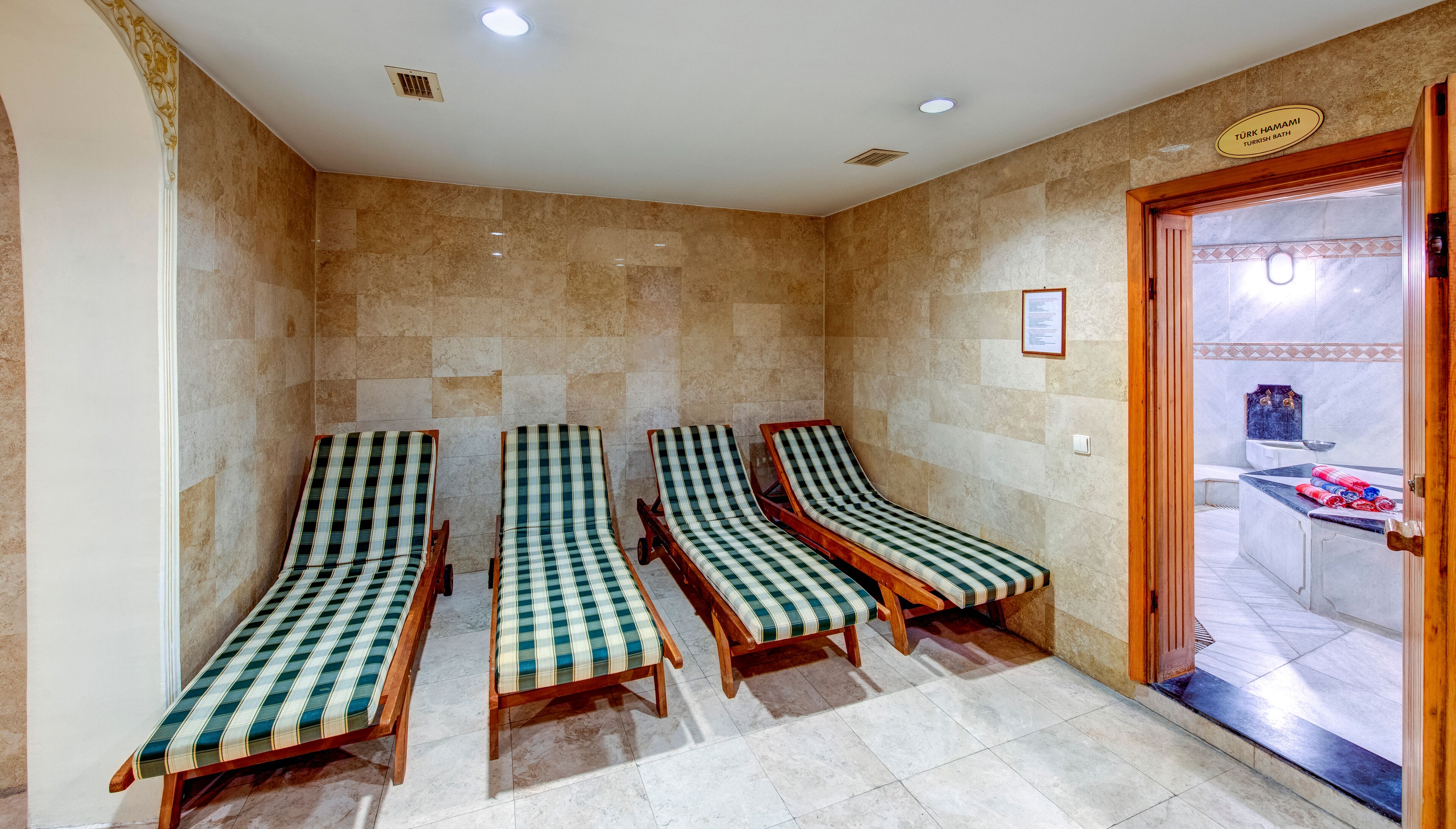 A Turkish bath with three wooden loungers, a tiled floor, and a door leading to a bathroom area.