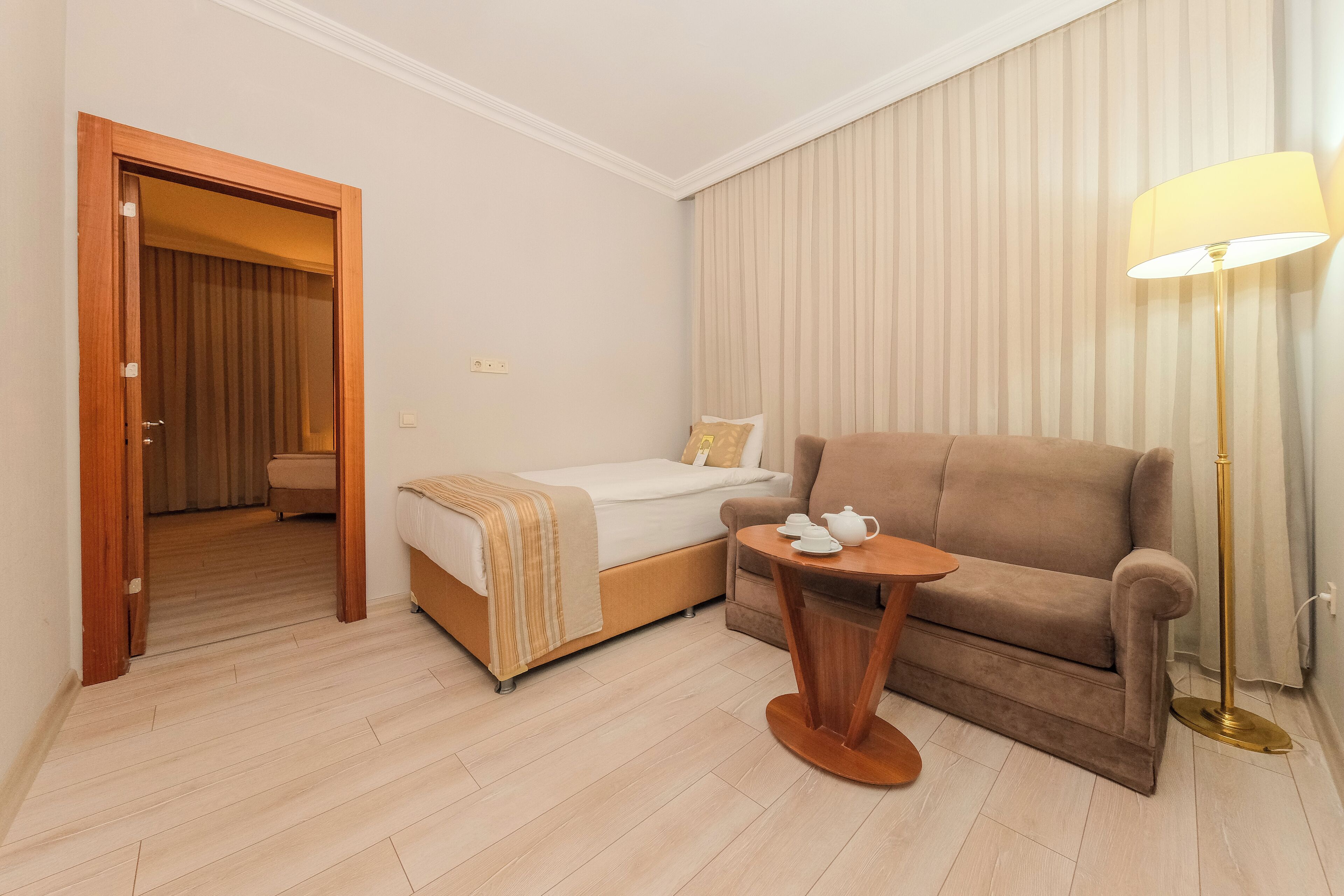 Habitación de hotel con cama, sofá, una mesita con lámpara y puerta que da a otra habitación.