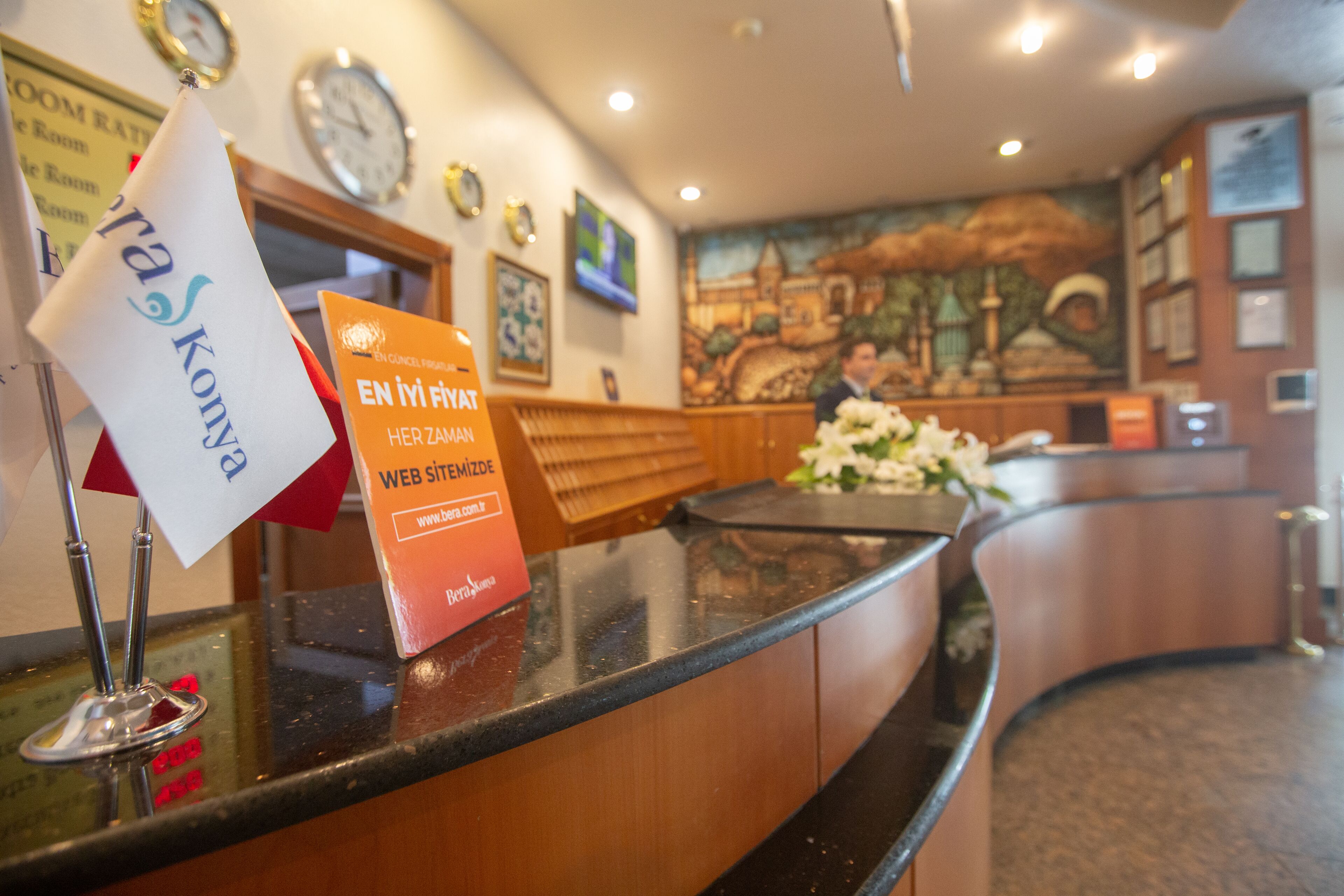 Un mostrador de recepción de hotel con un reloj, un cartel con tarifas de habitaciones y un arreglo floral.