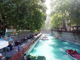 Un río con barcos, un muro de piedra y gente sentada en bancos a lo largo de la orilla.