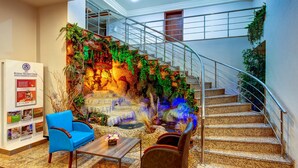 Vestíbulo de hotel con una escalera, un sillón azul, una mesa de madera y una pared con una cascada y follaje.