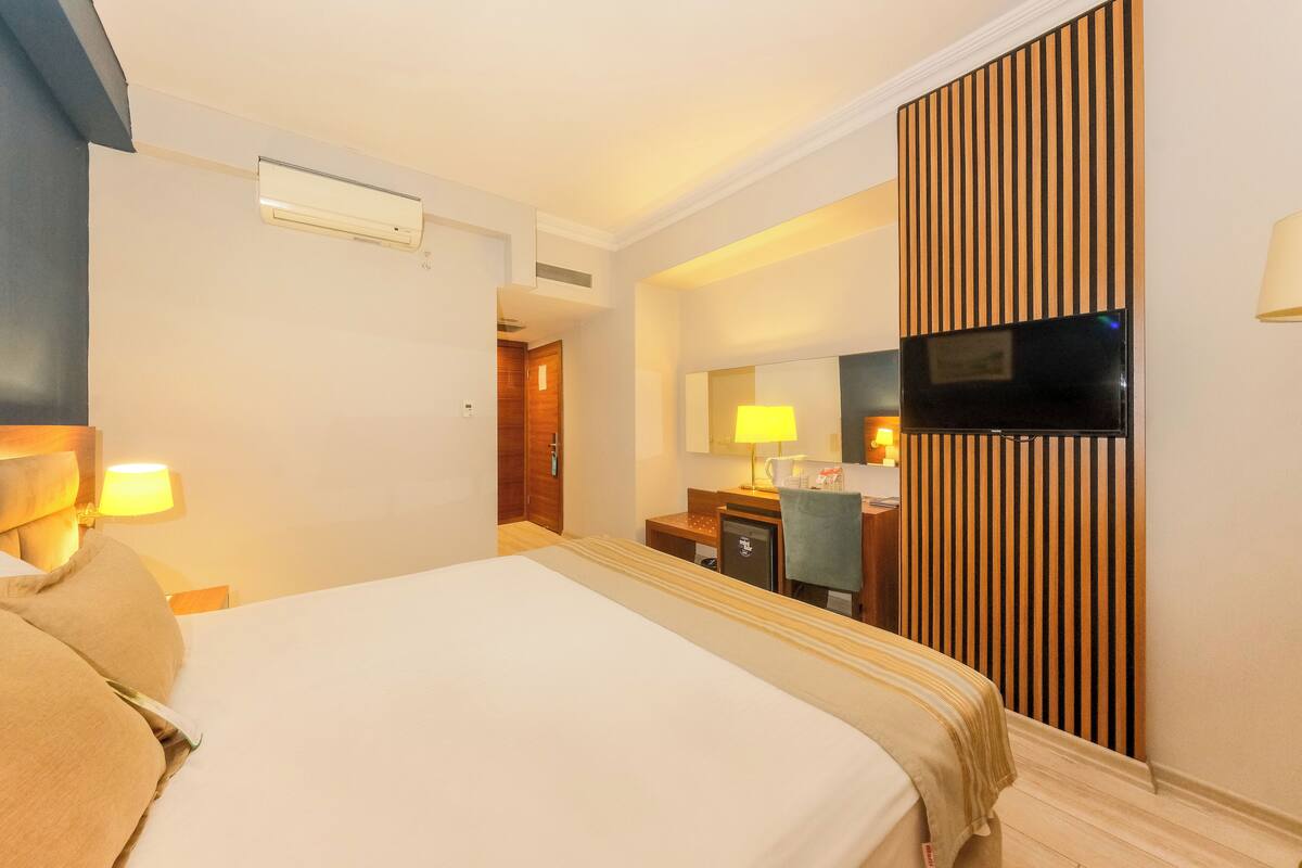 Una habitación de hotel con una cama, un escritorio con silla, un televisor y un aire acondicionado instalado en la pared.