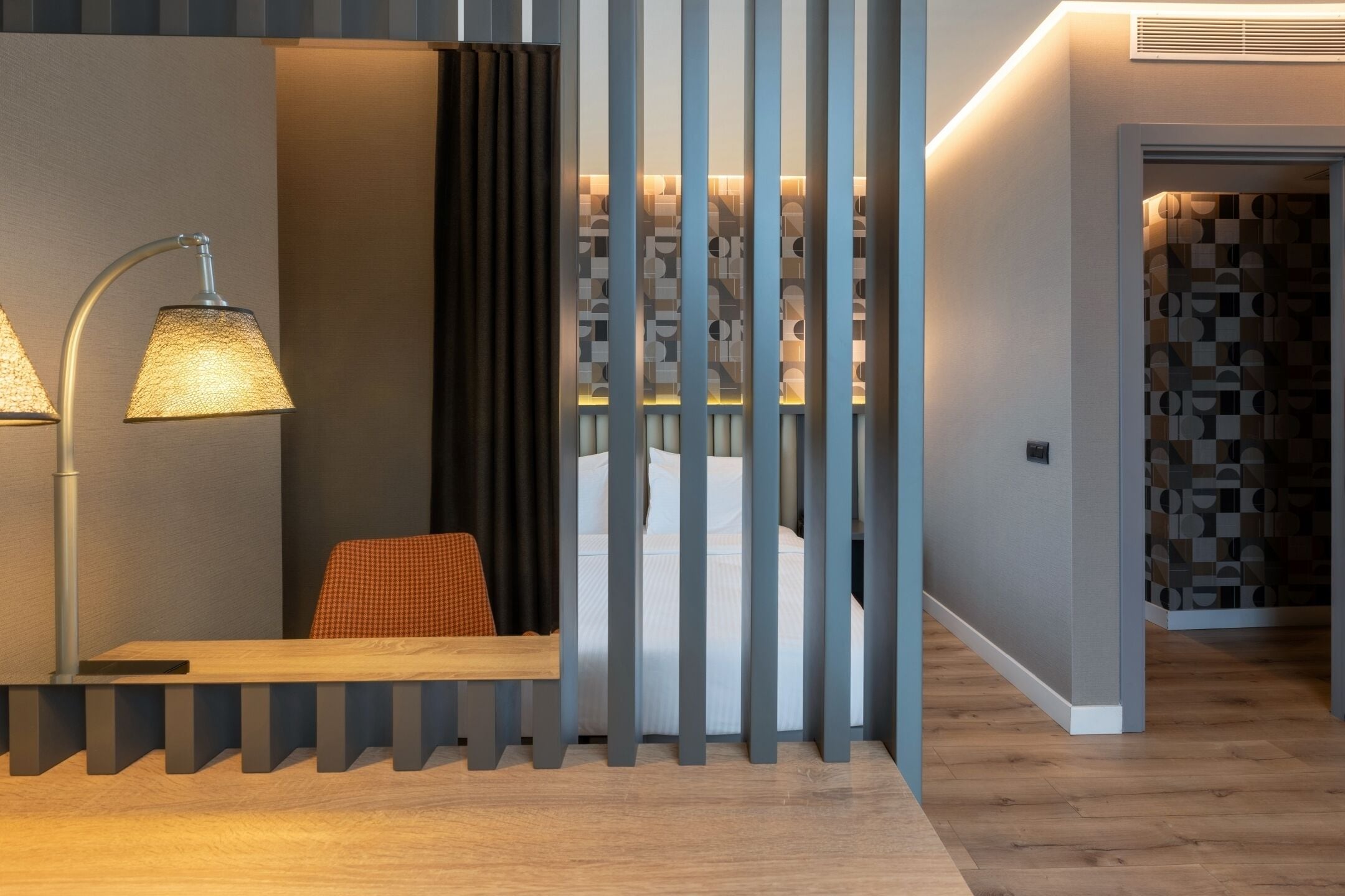 Een moderne hotelkamer met een houten vloer, een bureau met lamp, een bed met wit beddengoed en een kamerwand met patroon.
