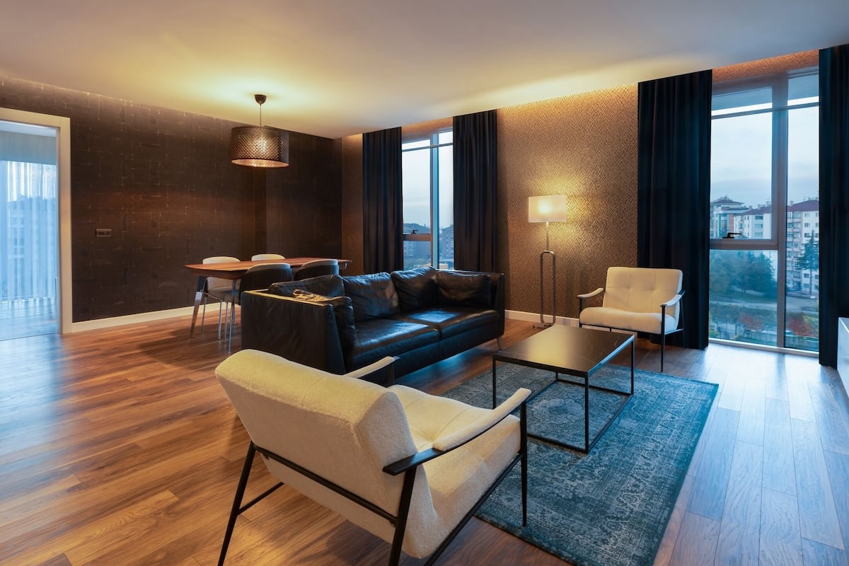 Un salón moderno con sofá, sillón, mesa de centro y comedor. Grandes ventanales ofrecen vista a la ciudad.