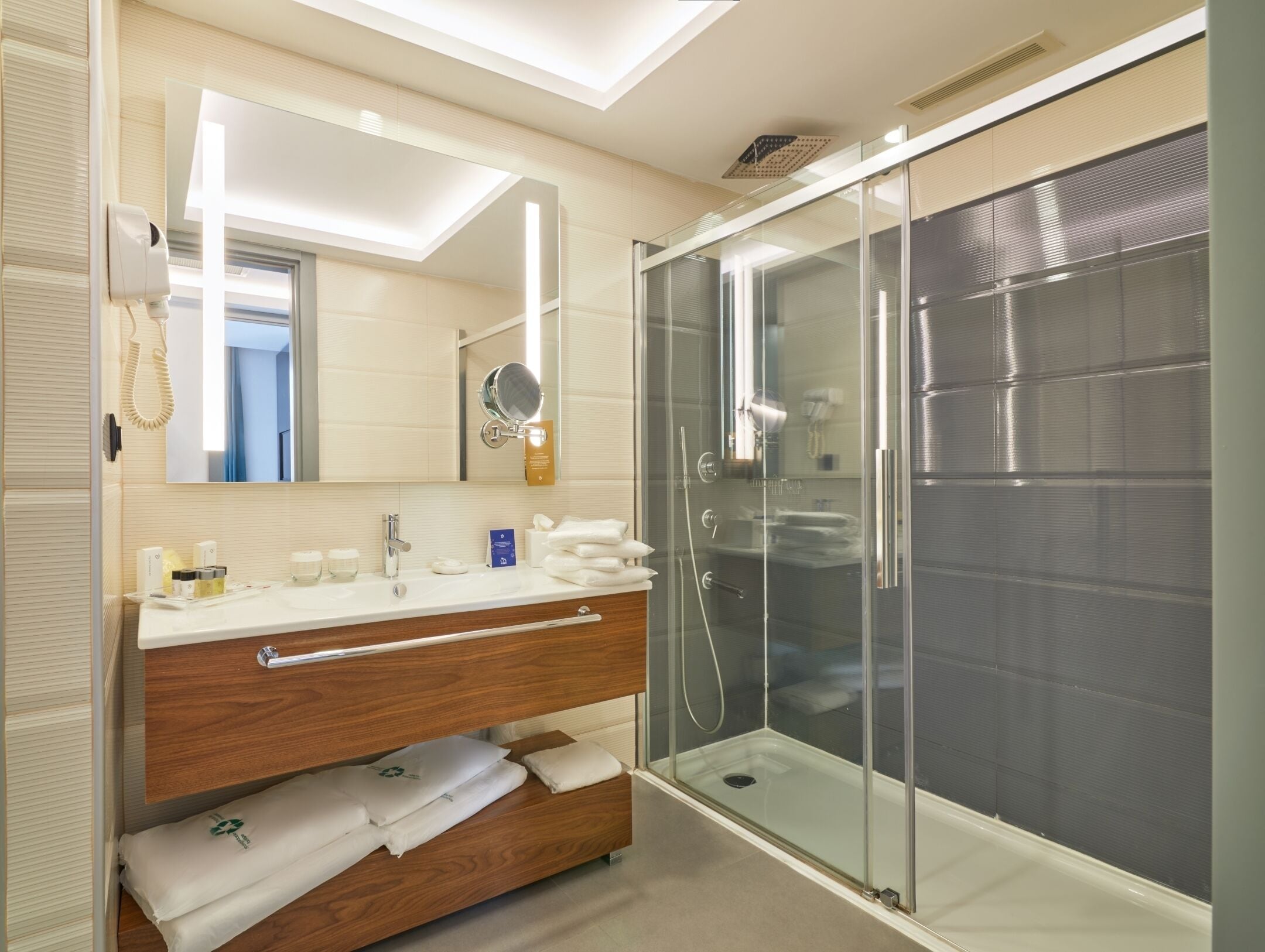A modern bathroom with a glass-enclosed shower, a vanity with a mirror, and neatly folded towels on a wooden shelf.