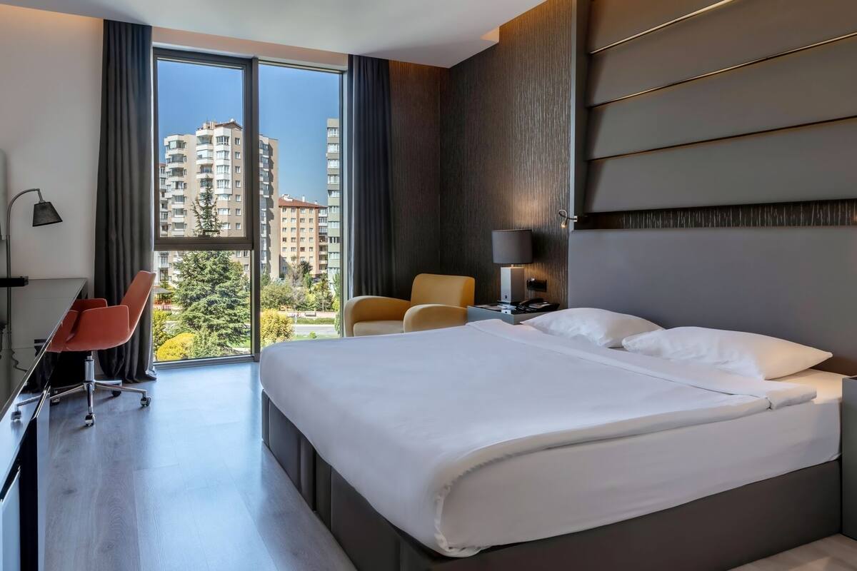 Habitación de hotel moderna con una cama grande, un escritorio con una silla, un sofá y vistas a rascacielos a través de un ventanal amplio.