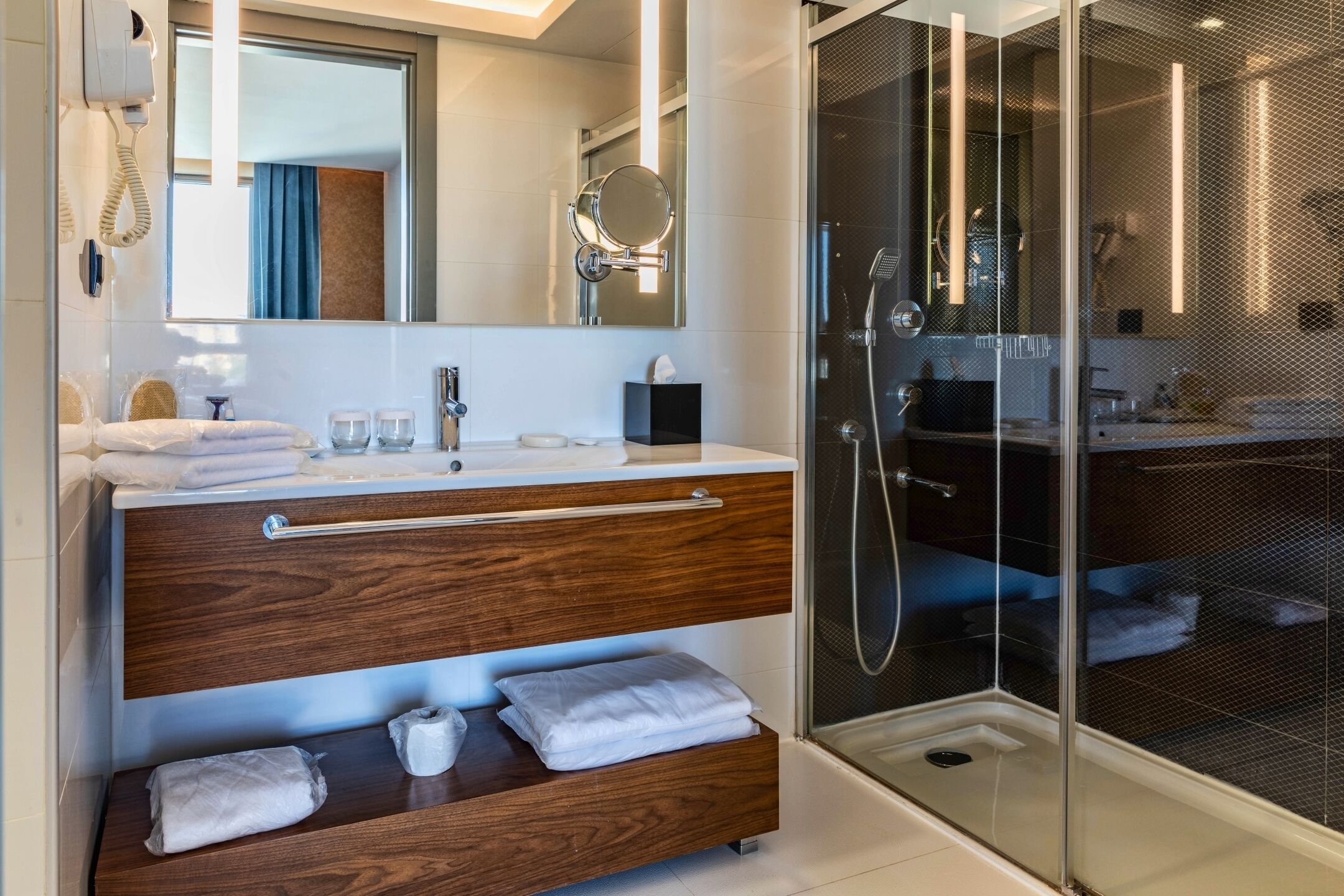 Un baño moderno con tocador de madera, un espejo grande, cabina de ducha de vidrio y un teléfono en la pared.