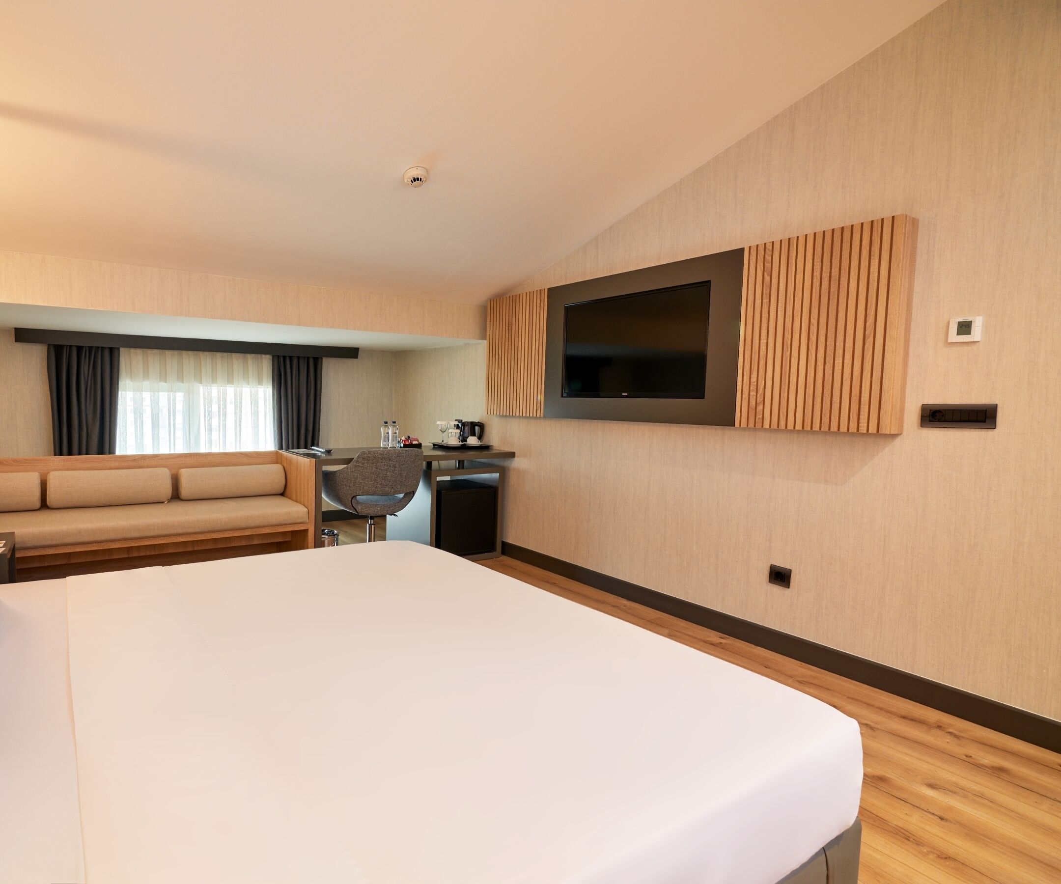 Une chambre d’hôtel moderne dotée d’un grand lit, d’un canapé, d’un bureau avec une chaise, d’un téléviseur à écran plat fixé au mur et d’une petite table avec une machine à café.