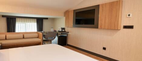 Habitación de hotel moderna con una cama grande, un sofá, un escritorio con una silla, un televisor de pantalla plana en la pared y una mesita con cafetera.