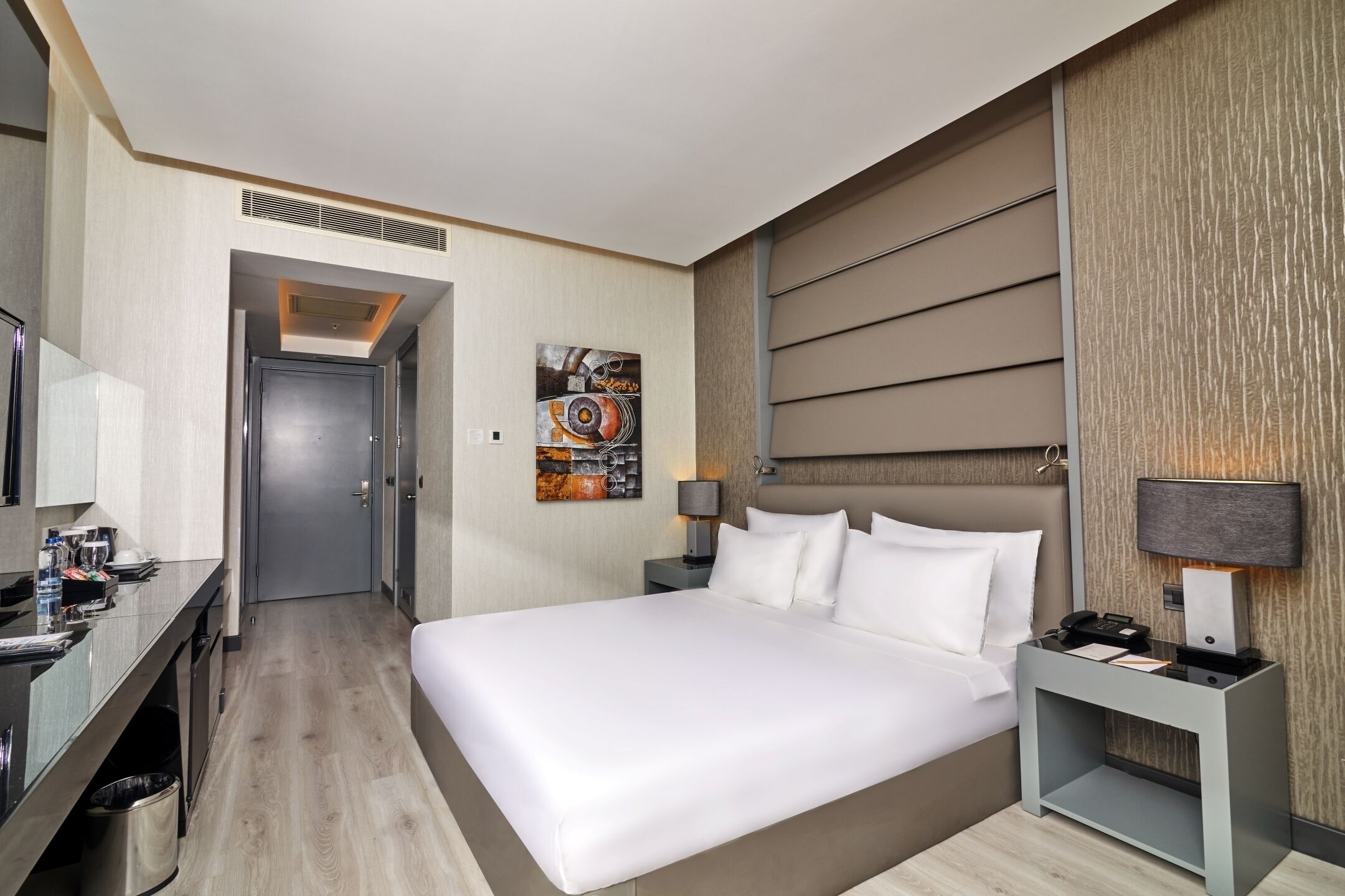 A modern hotel room with a large bed, a desk with a chair, a bedside table with a lamp, and a wall-mounted TV.