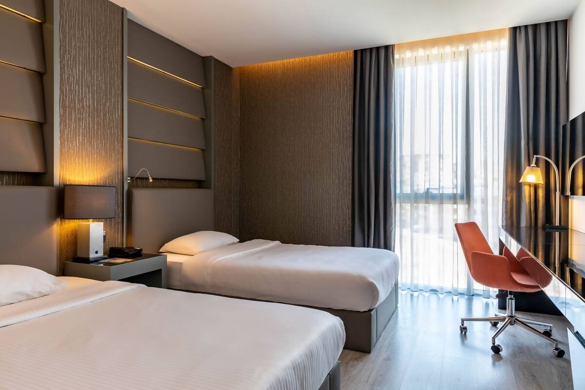 Habitación de hotel con dos camas, un escritorio, una silla y un ventanal con cortinas.