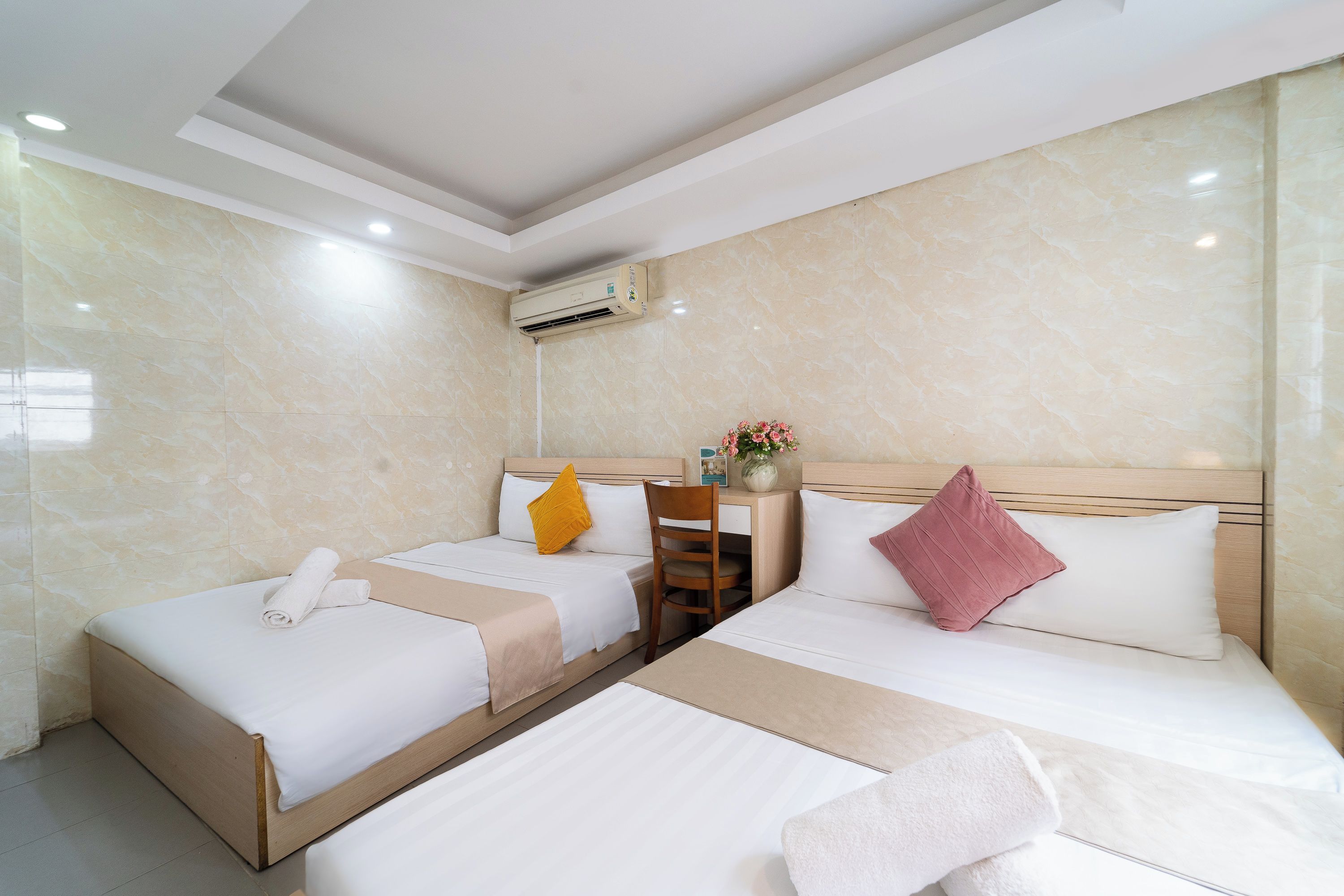 A hotel room with two beds, a desk, a chair, an air conditioning unit, and a vase with flowers.