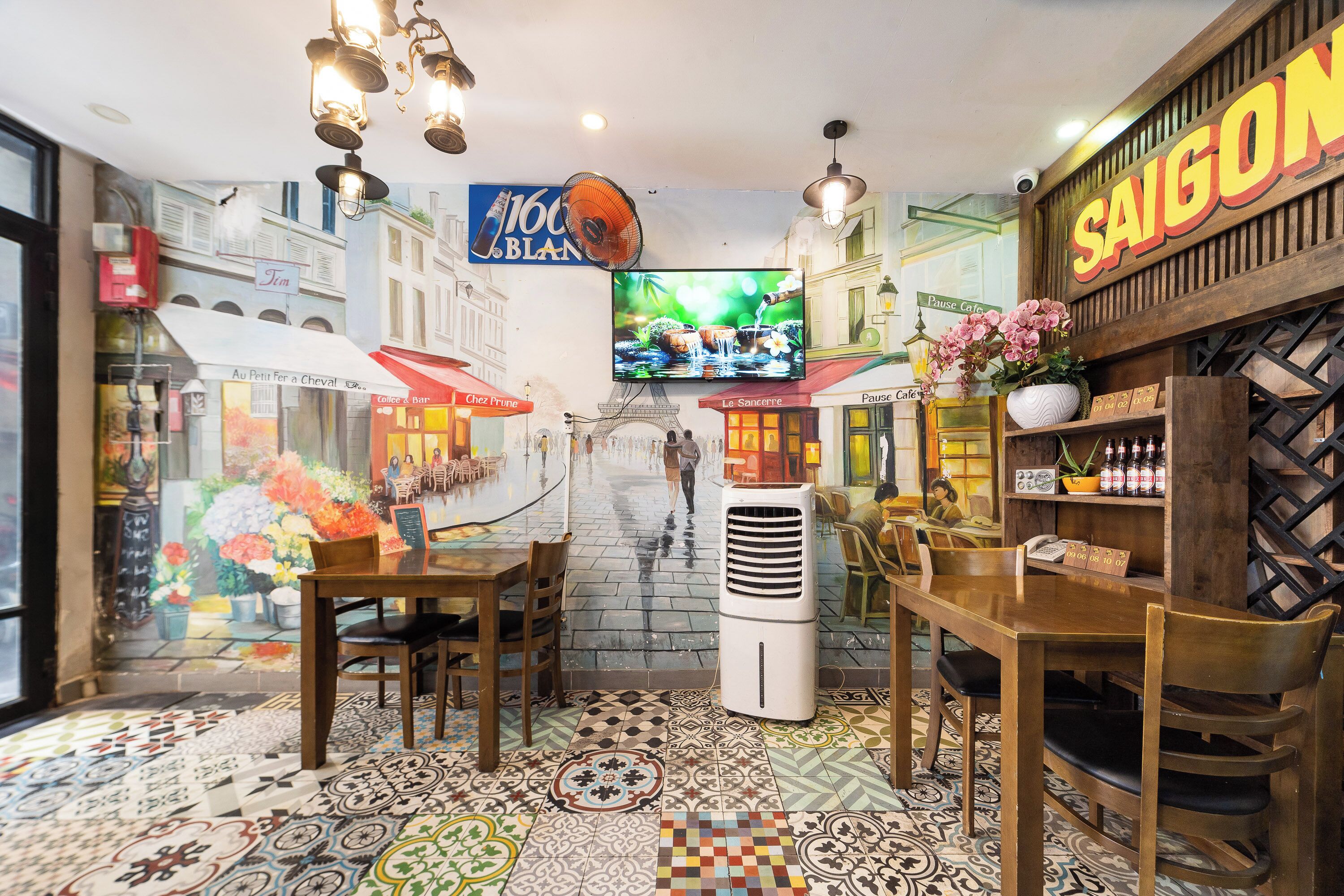 A restaurant interior with a mural of Parisian buildings, a television screen, and a variety of seating arrangements.