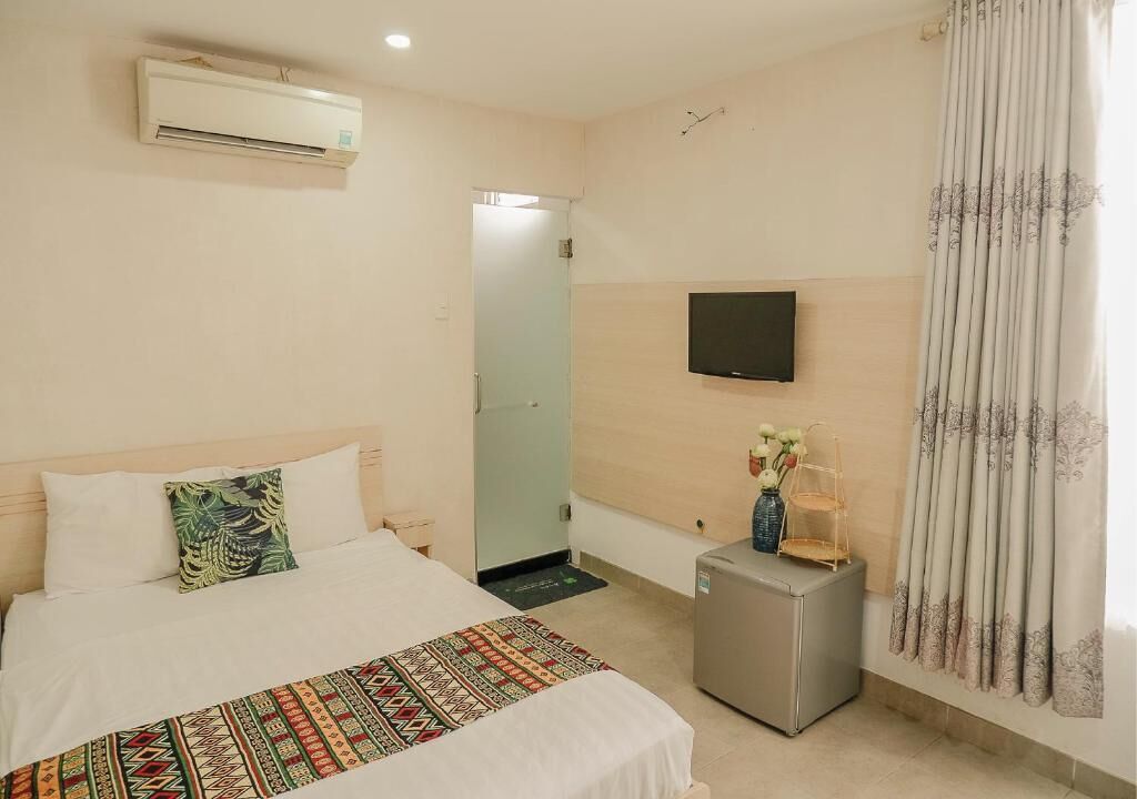A hotel room with a bed, a TV mounted on the wall, a small fridge, and a window with curtains.